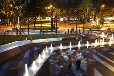 many people are walking around in the park at night with water fountains and lights on