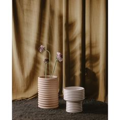 two white vases with flowers in them on a table next to a brown curtain