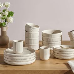 a bunch of white dishes and cups on a wooden table next to a vase with flowers