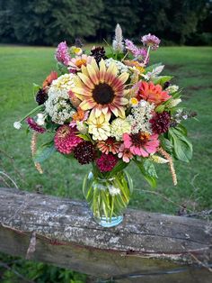 a vase filled with lots of flowers sitting on top of a wooden fence post in front of a field