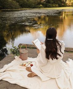 a woman sitting on a blanket reading a book near a lake with trees in the background