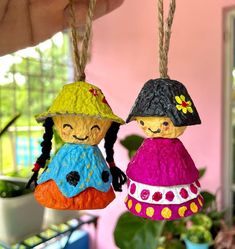 two little dolls hanging from twine strings in front of a potted plant and window