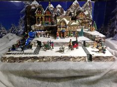 a christmas village is displayed in front of snow covered trees and mountains, with small figures on the ground