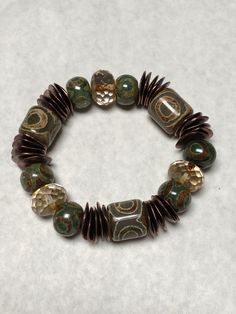 "PRITHVI (Earth Goddess):  This beautiful  bracelet seems so subtle in in its earthy coloring but the large (approx ½\" beads) take it to another level.  Subtle reddish tones to bronze wavy disc mykenos spacers.  Dzi design on barrel beads said to give spiritual protection.  Design includes transparent Picasso Premium Czech faceted rondelles  . . .  very large which \"airs out\" and lightens the pattern.   Strung on high quality pre-stretched elastic cord.  Rolls beautifully on the wrist.   I lo Large Beaded Bracelets, Big Bead Bracelets, Earthy Accessories, Earthy Crafts, Funky Bracelet, Earthy Jewelry, Earth Goddess, Earthy Green, Hippie Bracelets