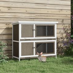 a rabbit sitting in the grass next to a wooden building with two cages on it