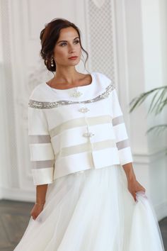 a woman wearing a white dress and jacket with silver accents on the shoulders, standing in front of a wall