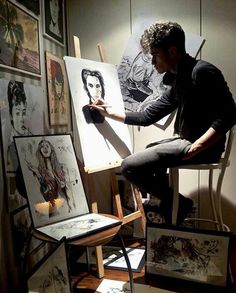 a man sitting in a chair drawing on an easel next to some pictures and paintings