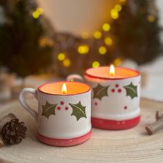 two mugs with holly designs on them sitting next to a pine cone and christmas tree