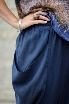 a woman with her hands on her hip wearing a blue skirt and gold ring, standing in front of a brick wall