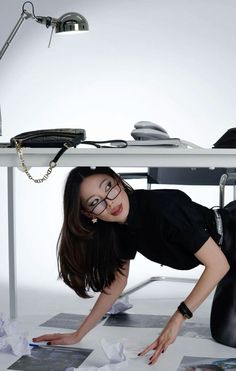 a woman in black shirt and glasses leaning over desk with papers on the floor next to her