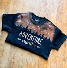 a t - shirt with the words adventure awaits on it sitting on a wooden floor