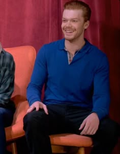 a man and woman sitting next to each other on orange chairs in front of a red curtain