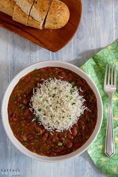 a white bowl filled with chili and rice next to a loaf of bread on top of a wooden cutting board