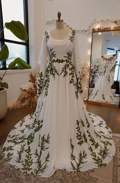 a white dress with green leaves is on display in front of a mirror and a potted plant