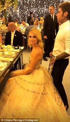 a woman in a ball gown sitting at a long table with people standing around it