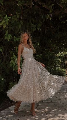 a woman in a white dress is standing on a stone path with trees and bushes behind her