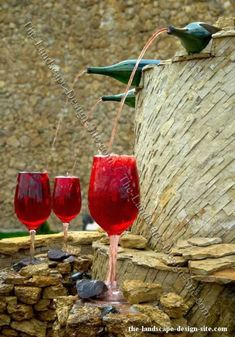 three red wine glasses are being filled with water from a fountain that has birds perched on top of it