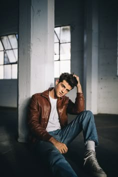 a young man sitting on the ground wearing a brown leather jacket and white t - shirt