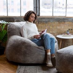 a woman sitting on a bean bag chair in a living room with large windows and potted plants