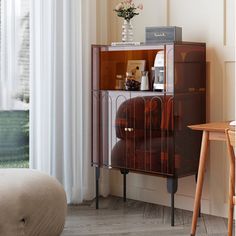 a brown cabinet sitting next to a window in a room with white curtains and a chair