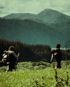 two people with backpacks are walking through the grass in front of some mountains and trees