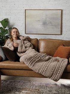 a woman is sitting on a couch with a blanket over her head and holding a cup