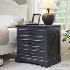 a night stand with two drawers and a lamp on it in front of a bed