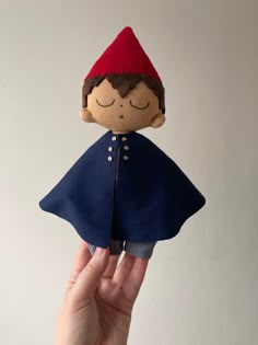 a hand holding a small doll with a red hat and blue cape on it's head