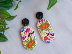 two earrings with designs on them sitting next to some plants