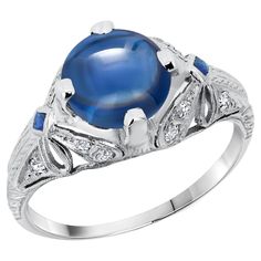 Indulge in timeless elegance with this exquisite vintage cocktail ring, a true masterpiece of fine jewelry craftsmanship. Featuring a stunning GIA Certified No Heat Burma Cabochon Round Sapphire, weighing an impressive 2.23 carats, this ring is a radiant symbol of sophistication and luxury. The captivating sapphire is set in a beautifully engraved and millgrain platinum band, exuding a vintage charm that is both unique and enduring. Main Features: Center Stone: Type: GIA Certified No Heat Sapphire Shape: Cabochon Round Weight: 2.23 Carats Color: Deep, Rich Blue Accent Stones: Diamonds: Total Weight: 0.15 Carats Cut: Round Brilliant Setting: Pave, enhancing the ring’s sparkle Side Sapphires: Shape: Square Color: Matching deep blue, perfectly complementing the center stone Ring Setting: Meta Luxury Sapphire Ring With Cabochon, Yellow Sapphire Rings, Vintage Cocktail Ring, Round Sapphire, Platinum Diamond Rings, Blue Accent, Gold Cocktail Ring, Diamond Cocktail Rings, Sapphire Diamond Ring