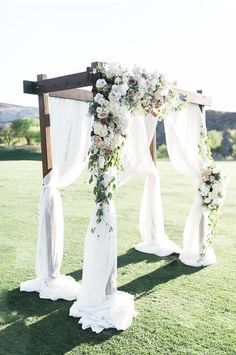 an outdoor wedding ceremony with white flowers and draping on the arbor, in front of a green field