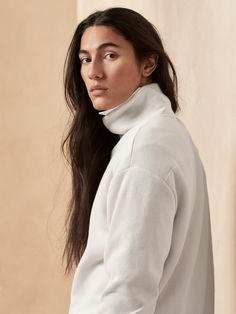 a woman with long hair wearing a white turtle neck sweater and jeans, standing in front of a beige wall