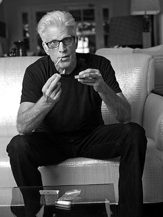 an older man sitting on a couch eating something with his hands and looking at the camera