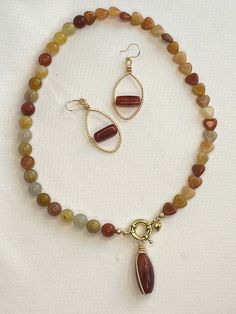 Conjunto de joyería elegante y natural compuesto por un collar y un par de aretes. El collar está elaborado con cuentas suaves y redondeadas en tonos cálidos, incluyendo matices de marrón, rojo y beige. Destaca un cierre dorado con un diseño detallado, del cual cuelga un colgante de piedra alargada en color rojo, envuelto en delicado alambre dorado. Los aretes combinan perfectamente con el collar, cada uno con una piedra roja alargada colocada horizontalmente en el centro de un marco ovalado retorcido en color dorado. Cuentan con ganchos dorados sencillos para un ajuste cómodo. Este conjunto es ideal para quienes buscan una apariencia elegante y terrenal, con detalles en piedras naturales y toques dorados que crean una armonía perfecta. Este conjunto es creado por una pequeña empresa propi Color Rojo, Color Dorado, Pendant Necklaces, Labour Day, Jewelry Necklace Pendant, Beauty Book, Accessory Gift, Jewelry Necklaces, Display Homes