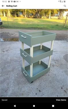 a green cart with three drawers sitting on top of cement ground next to grass and trees