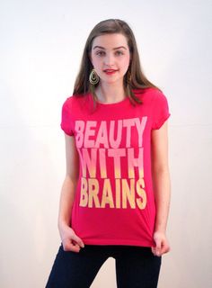 This is a soft 90s vintage hot pink women's t-shirt with cap sleeves and large letter graphics on the front in a red grapefruit color and yellow, which spell out "Beauty With Brains." The letters are outlined in metallic silver. Show the world that you are not just a pretty face! Lightweight and super soft and broken in, it makes a great workout tee.The shirt is in good, clean condition with very minor wear on some of the silver metallic edges and some subtle variations in the pink color on the Vintage Pink T-shirt With Graphic Design, Vintage Pink T-shirt With Funny Print, Unisex Pink T-shirt With Funny Print, Fun Graphic Design Tops, Fun Pink Top With Graphic Design, Spring Pink Pop Culture Tops, Pink Pop Culture Tops For Spring, Pink Pop Culture Tops With Funny Print, Pink Pop Culture Top With Funny Print