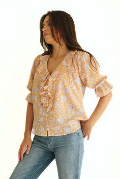 Discover our Chloe Shirt. Ethically made from a sustainable soft cotton orange and white floral print, she exudes comfort and style. Designed as a voluminous blouse, she features timeless, feminine details such as a ruffle neckline, real shell buttons, and petit pin-tucks. A true closet staple, the Chloe Shirt can be styled from day to night all year long. Responsibly Handcrafted by women artisans using high-quality sustainable fabrics certified Oeko Tex Standard 100.1% of your order funds regen Cotton Ruffle Sleeve Tops With Floral Print, Cotton Top With Floral Print And Ruffle Sleeves, Cotton Tops With Floral Print And Ruffle Sleeves, Cotton Tops With Ruffle Sleeves And Floral Print, Cotton Blouse With Floral Print And Ruffle Sleeves, Feminine Cotton Blouse With Ditsy Floral Print, Cotton Floral Print Top With Ruffled Collar, Cotton Tops With Floral Print And Ruffled Collar, Printed Cotton Blouse With Ruffle Sleeves