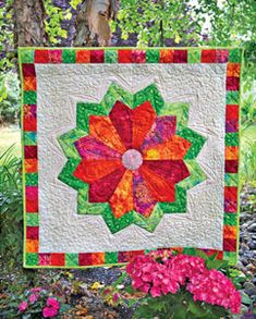 a quilted wall hanging on the side of a tree with flowers in front of it