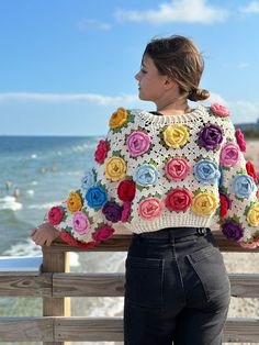 a woman standing on the beach wearing a colorful crochet sweater with roses in it