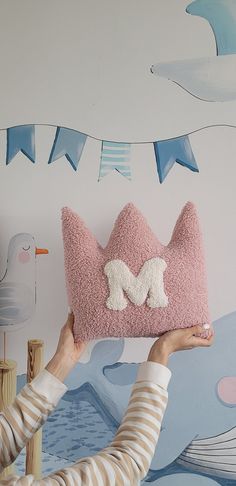 a woman holding up a pink pillow with the letter m on it in front of a wall