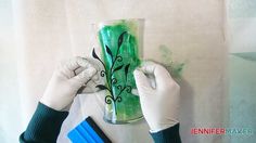 two hands in white gloves painting a vase with green and black designs on the side