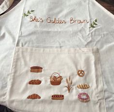 a white apron with embroidered images of bread and pastries on the front, along with words that read ski's golden brown