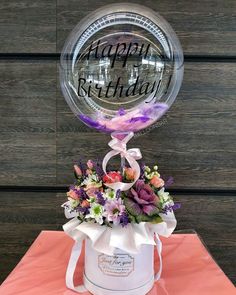 a happy birthday balloon sitting on top of a white bucket filled with flowers and greenery