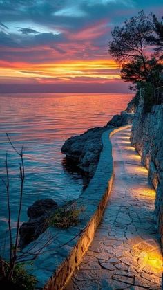 a path leading to the ocean at sunset with lights on it and trees growing out of the rocks