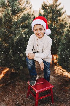 a young boy in a santa hat sitting on a small red chair near christmas trees