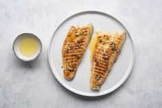 two fish fillets on a white plate next to a small bowl and sauce container