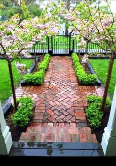 a brick walkway with trees and flowers on either side, leading to an open gate