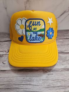 Lake Day Patch Trucker Hat The front of this hat is embellished with patches including one that says fun at the lake, flowers and a heart. This hat is perfect for a day out at the lake or day on the boat. The blue and yellow colors are perfect for summer! The hat has a foam front panel and a mesh back with and adjustable strap in the back. Fun Spring Hats For Outdoor Activities, Fun Spring Outdoor Hats, Spring Outdoor Trucker Snapback Hat, Yellow Trucker Hat Baseball Cap For Spring, Spring Snapback Trucker Hat For Outdoor Activities, Yellow Trucker Hat With Curved Brim For Spring, Retro Yellow Hats For Outdoor, Retro Yellow Hat For Outdoor, Retro Yellow Outdoor Hat