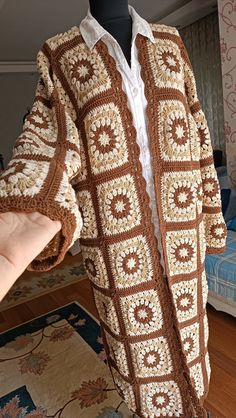 a crocheted jacket is being held up by a person's hand in front of a bed