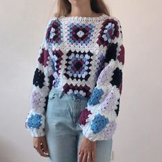 a woman wearing a crocheted sweater and jeans stands in front of a white wall